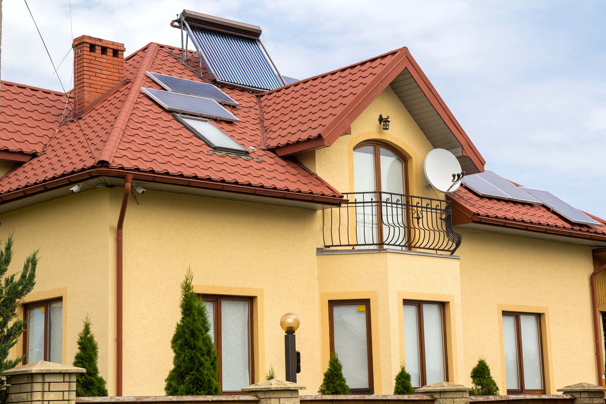 Close-up of new brick house top with red shingle roof, satellite dish, solar panels and plastic attic windows on bright blue sky background. Real estate property and professionally done work concept
